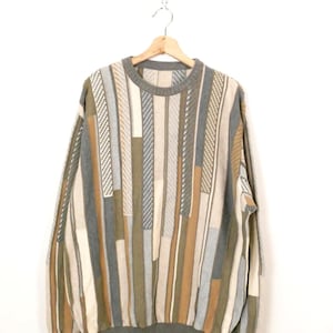 Puede incluir: Un su&eacute;ter vintage de cuello redondo con un patr&oacute;n de rayas verticales en tonos beige, marr&oacute;n, gris y verde oliva. El su&eacute;ter tiene cuello, pu&ntilde;os y dobladillo de canal&eacute;. La prenda est&aacute; colgada en una percha de madera.