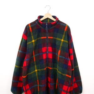 May include: A fleece pullover with a tartan pattern in red, green, blue, and yellow. The long-sleeved top has a quarter-zip closure at the neck and a small zipped pocket on each side. The garment is hanging on a wooden hanger.