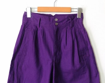 Puede incluir: Pantalones cortos morados de cintura alta con pliegues y cierre de dos botones. Los pantalones cortos cuelgan de una percha de madera. Tienen dos bolsillos delanteros. Fondo blanco.