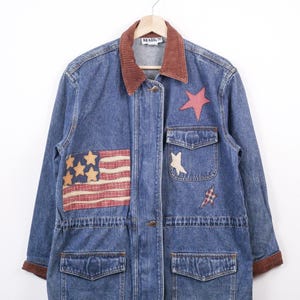 May include: Blue denim jacket with a brown corduroy collar and cuffs. The jacket features patches of the American flag, stars, and a plaid star. It has four front pockets and a button-up closure.