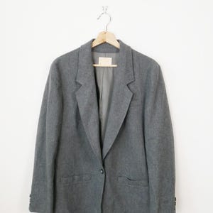 May include: A grey blazer hanging on a wooden hanger against a white wall. The blazer features a single-button closure, notched lapels, and two front pockets. The fabric appears to be a wool blend.