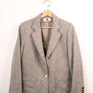 Peut inclure: Un blazer gris clair chiné doté d'une fermeture à un bouton, de revers crantés et de deux poches plaquées à l'avant. Les manches présentent trois petits boutons noirs aux poignets. Une étiquette blanche avec le texte bleu EJE est au col.