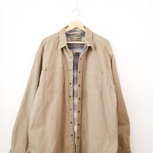 Op de afbeelding: Een beige overhemd met lange mouwen, knoopsluiting, kraag en twee borstzakken. De binnenvoering heeft een blauw, bruin en crèmekleurig ruitpatroon. Het label bij de nek vermeldt L.L.Bean en het overhemd hangt aan een houten kledinghanger.