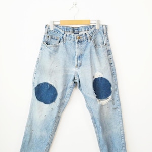 May include: Light-wash blue denim jeans featuring a classic five-pocket design, a button fly, and two prominent circular dark blue fabric patches sewn onto the knees. The denim shows signs of wear with light distressing and scattered dark spots.