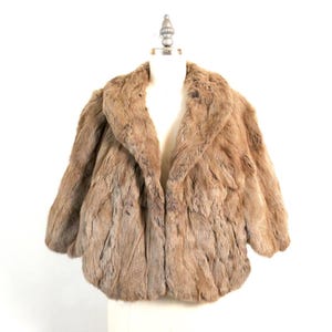 Peut inclure: Une veste en fourrure brun clair dotée d'un large col châle, de manches trois-quarts et d'une coupe courte. Le vêtement est présenté sur un mannequin, mettant en valeur la matière en fourrure dense et texturée ainsi que le design ouvert.