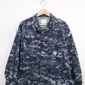 Può includere: Giacca mimetica digitale blu navy e grigia con chiusura a bottoni, due tasche sul petto e una piccola toppa sulla tasca destra. La giacca è appesa a una gruccia di legno su sfondo bianco.
