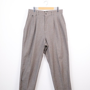 May include: Brown and gray patterned trousers hanging on a wooden hanger. The pants have a button and zipper closure, belt loops, and pleats at the waist. The fabric has a subtle checkered design.