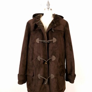 May include: A brown duffel coat with a hood and toggle closures. The coat has a faux suede exterior and a warm, fur-like lining. Features include two front pockets and a classic, timeless design.