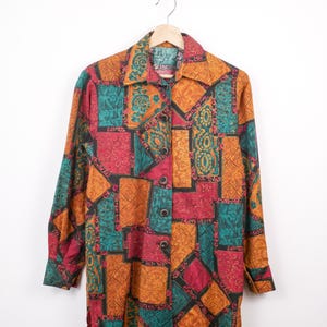 May include: A long-sleeved shirt with a patchwork design in orange, teal, red, and black. The shirt is hanging on a wooden hanger against a white background. The pattern features geometric and floral motifs. The shirt has a collared neckline.