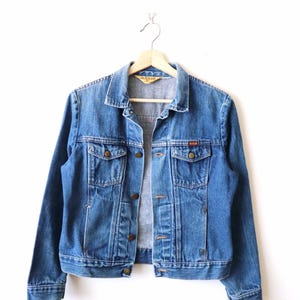 May include: A medium-wash blue denim jacket featuring a classic collar, long sleeves, and a button-front closure. The garment includes two chest pockets with buttoned flaps and metal hardware. A yellow brand label is visible inside the neck area.