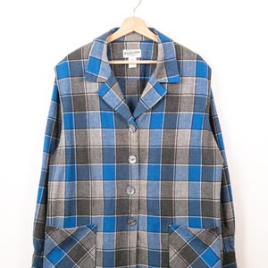 May include: A long-sleeve button-down shirt featuring a blue, grey, and black plaid pattern. The shirt has a notched collar, four front buttons, and two square patch pockets at the bottom. A Pendleton brand label is visible at the inner neckline.