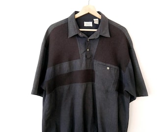 May include: A short-sleeve polo shirt in shades of charcoal grey and black. The shirt features a collared neckline with a three-button placket, a single chest pocket with a button closure, and a solid black ribbed hem at the bottom of the garment.
