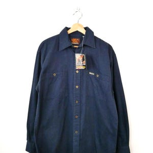 以下が含まれることがあります： 木製のハンガーに掛けられた、ボタンダウンの濃紺のシャツ。胸ポケットが2つと襟が付いています。シャツには「Grizzly」と書かれたタグが付いています。柔らかく丈夫な生地で作られています。