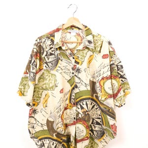 May include: Cream-colored button-down shirt with a map and compass design. The short-sleeved shirt features a repeating pattern of vintage maps, compasses, and nautical elements in shades of green, brown, and yellow.