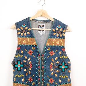 May include: A blue denim vest featuring intricate embroidery with floral and geometric patterns in red, yellow, and turquoise. The garment includes brown fabric bands at the shoulders and hem, with a label reading ROPA and size M at the neck.