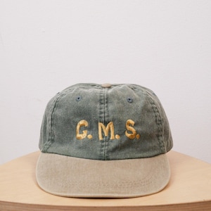May include: A baseball cap featuring a faded olive green crown and a contrasting tan brim. The front of the cap displays the letters G.M.S. embroidered in gold thread. The cap includes a small tan button at the top and matching eyelets.