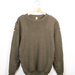 May include: An olive green knit sweater featuring a crew neckline, ribbed cuffs, and reinforced fabric patches on the shoulders. Small German flag patches are stitched onto the upper sleeves of this long-sleeved pullover garment.