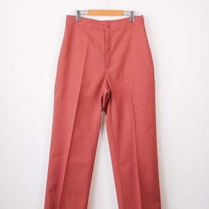 May include: Rust-coloured high-waisted trousers hanging on a wooden hanger. The trousers have a button closure and a straight-leg design. The fabric appears to be a medium-weight material, suitable for various seasons.