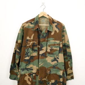 Peut inclure: Veste de camouflage avec plusieurs poches et le texte "USMC" sur la poitrine. La veste est dans des tons de vert, marron et noir, suspendue à un cintre en bois sur fond blanc.