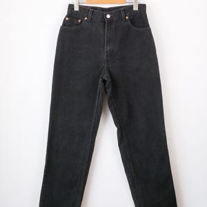 Op de afbeelding: High-waisted zwarte spijkerbroek van denim met een klassiek ontwerp met vijf zakken, een metalen knoopsluiting, een ritssluiting en koperkleurige klinknagels. De broek heeft een rechte pijp en hangt aan een houten kledinghanger.