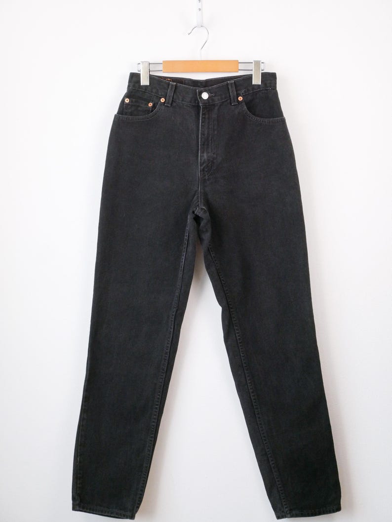 Op de afbeelding: High-waisted zwarte spijkerbroek van denim met een klassiek ontwerp met vijf zakken, een metalen knoopsluiting, een ritssluiting en koperkleurige klinknagels. De broek heeft een rechte pijp en hangt aan een houten kledinghanger.