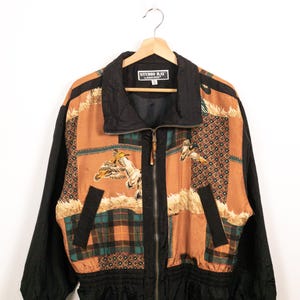 May include: A Studio Ray zip-up jacket featuring a patchwork design with black sleeves, a black collar, and a central panel displaying ducks in flight, plaid patterns, and geometric motifs in shades of brown, green, and tan with a black waistband.