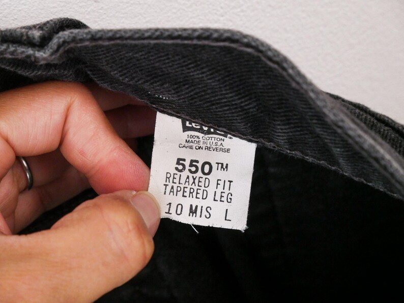 Op de afbeelding: Een zwart denim kledingstuk met een wit binnenlabel met de tekst Levi's, 100% cotton, Made in U.S.A., Care on reverse, 550 Relaxed Fit Tapered Leg en 10 MIS L. De stof heeft een zichtbare keperbinding en donkergrijze tinten.