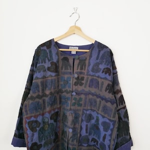May include: A vintage button-down jacket with a distinctive patchwork design. The jacket combines blue, grey, and brown hues, showcasing repeating patterns of elephants and floral motifs. It has long sleeves and a round neckline.