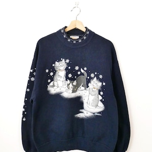 Könnte beinhalten: Ein marineblaues Langarm-Sweatshirt mit einer Grafik von drei Kätzchen, die im Schnee spielen. Das Design umfasst weiße Schneeflocken, Pfotenabdrücke und einen gemusterten Kragen. Die Kätzchen sind in Grau-, Schwarz- und Weißtönen dargestellt.
