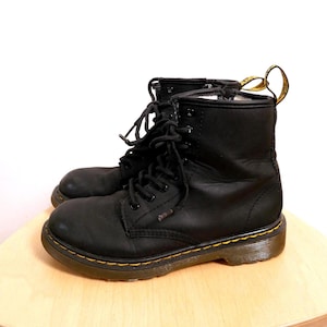 Peut inclure: Une paire de bottes de combat en cuir noir avec des lacets noirs et des coutures jaunes autour des semelles. Les bottes sont sur une surface en bois brun clair. Les bottes ont un design classique et robuste.