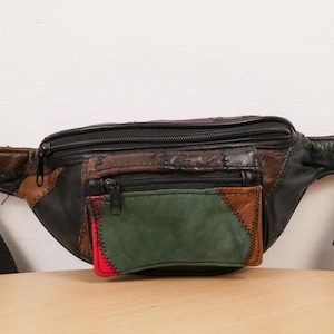 Peut inclure: Une sacoche banane en cuir multicolore présentant un motif patchwork dans des tons de noir, marron, vert foncé et un petit panneau rouge. Le sac comprend un compartiment principal zippé et une poche avant zippée plus petite avec des sangles noires.
