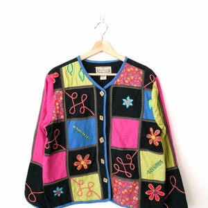 Puede incluir: Una chaqueta negra de botones con un diseño de patchwork que presenta cuadrados coloridos con flores, hojas y remolinos bordados. La chaqueta tiene un cierre de botones y mangas largas.
