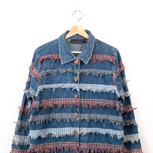 Peut inclure: Une veste en jean bleu avec des bandes horizontales de tissu à carreaux rouges et bleus. La veste présente un col, des manches longues et une fermeture à cinq boutons marron. Chaque panneau à carreaux est agrémenté de bords effilochés.