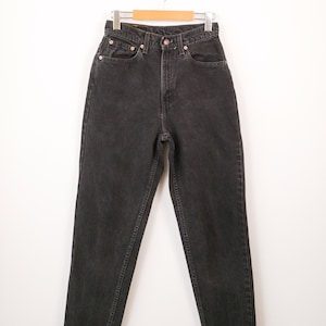 May include: High-waisted black denim jeans featuring a classic five-pocket design, a button fly closure, and copper-toned metal rivets. The jeans have a straight-leg cut with visible stitching along the seams and are displayed on a wooden hanger.