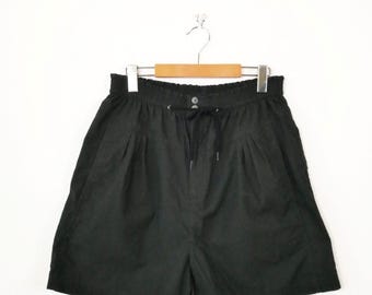 Puede incluir: Pantalones cortos negros con cintura elástica y cordón ajustable, dos botones frontales y detalles de pliegues en la parte delantera. Los pantalones están colgados en una percha de madera con gancho metálico sobre fondo blanco.