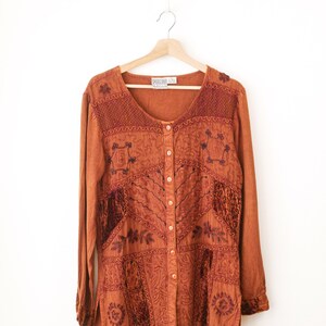 May include: A long-sleeved, button-down shirt in a burnt orange hue, featuring intricate dark brown embroidery and textured velvet-like panels. The garment has a round neckline and a straight hem, displayed on a wooden hanger against a white background.