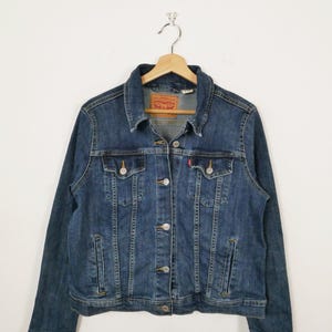 Op de afbeelding: Een blauw spijkerjack met een klassiek ontwerp, met een knoopsluiting, borstzakken en een kraag. De jas hangt aan een houten hanger tegen een witte achtergrond. Een rood en wit label is zichtbaar op de jas.