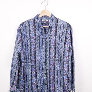 May include: A patterned button-down shirt in shades of blue, black, and purple. The long-sleeved shirt is displayed on a wooden hanger. The shirt features a paisley design and a collar, with buttons down the front.