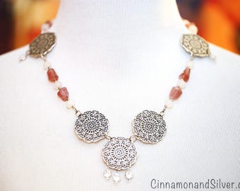 May include: A necklace featuring five circular silver-toned filigree medallions connected by strands of translucent white beads and faceted reddish-brown stones. The central medallion has three small dangling beads at the bottom edge.