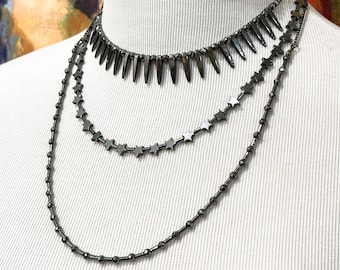 May include: Three-strand necklace set. The top necklace features dark gray spikes, the middle one has star-shaped beads, and the bottom one has small round beads. The necklaces are displayed on a white mannequin.