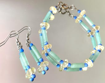 May include: A matching jewelry set featuring earrings and a bracelet. The earrings have silver hooks and are adorned with blue, light green, and yellow beads. The bracelet has a double strand design with similar beads.