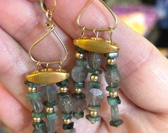 May include: A pair of dangle earrings featuring gold-tone wire hooks and triangular frames. Each earring has a horizontal gold-tone bar suspending three strands of alternating small green stone chips, translucent grey faceted beads, and tiny gold-tone metal spheres.