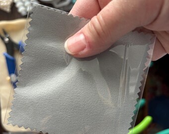 May include: A gray microfiber cleaning cloth in a clear plastic package. The cloth has a textured surface and a zig-zag edge. The background includes jewelry-making supplies, suggesting a product for cleaning jewelry or other delicate items.