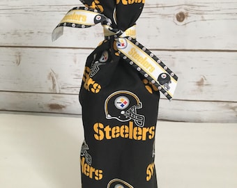 May include: Black fabric wine bottle bag featuring the Pittsburgh Steelers logo and the word "Steelers" in gold. The bag is tied with a white ribbon with the team's name and logo. A decorative gift bag.