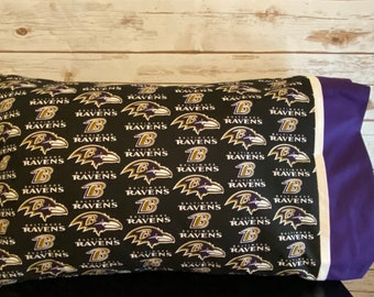 May include: A black and purple pillow with a repeating pattern of the Baltimore Ravens logo. The logo features a raven with the words "Baltimore Ravens" and a "B" in gold.