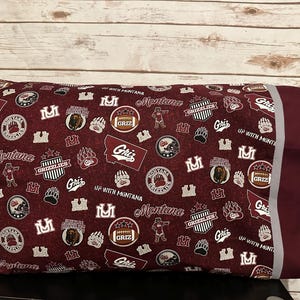 May include: A maroon pillowcase with a white and gray trim. The pillowcase features a repeating pattern of the University of Montana Grizzlies logo, the words "Montana" and "Griz", and other related imagery.