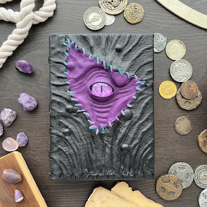 May include: A black leather journal with a purple eye design stitched onto the cover with teal thread. The eye is centered on the cover and surrounded by a textured, dark gray background. Coins and gemstones are scattered around the journal.