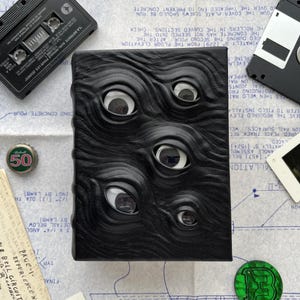 Könnte beinhalten: Ein schwarzer, strukturierter Notizbuchdeckel mit mehreren weißen Augendesigns. Das Notizbuch ist von einer Kassette, einer Diskette, einem Dia und einem Flaschenverschluss umgeben. Das Design des Notizbuchs verleiht ihm ein einzigartiges, künstlerisches Aussehen.