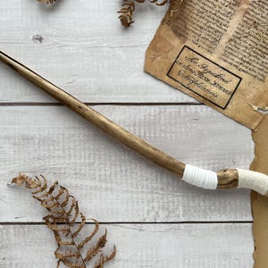 May include: A handcrafted wooden wand with a pointed tip and a wrapped handle. The wand is light brown and has a natural wood grain. The handle is wrapped with white fabric. The wand is lying on a white wooden surface with dried leaves and aged paper.