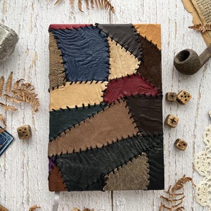 May include: A handcrafted journal featuring a patchwork leather cover in various colours, including blue, tan, brown, and burgundy, stitched with black thread. The journal rests on a white wooden surface, surrounded by vintage items.
